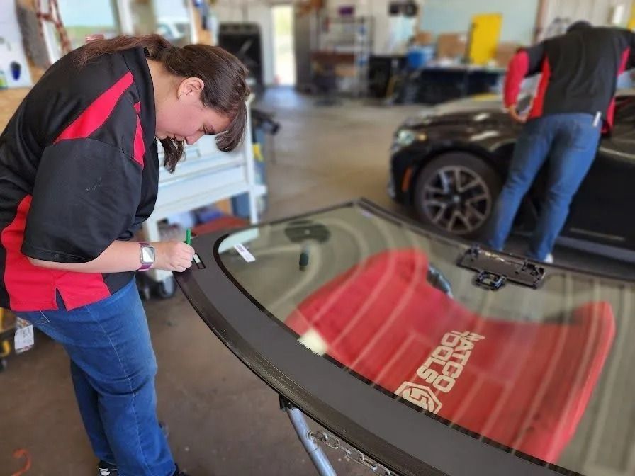 A worker in a black and red uniform works on a car windshield in an auto repair shop while another person works on a car.