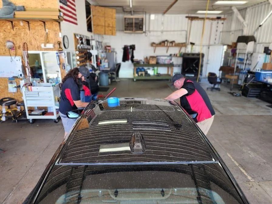 Two technicians in matching red and black shirts work on the roof of a vehicle inside an auto repair shop.