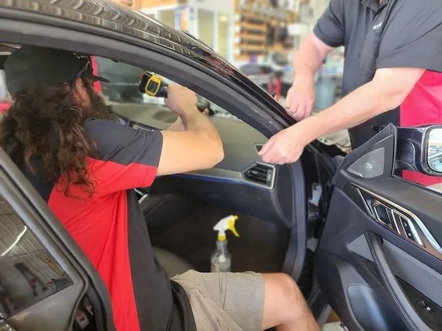 Two auto shop employees work on the interior of a vehicle, with one holding a tool near the dashboard.