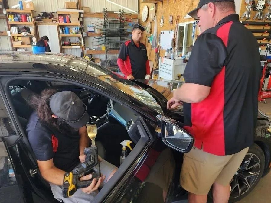Two workers in a shop install window tint on a car, with a third person working in the background.