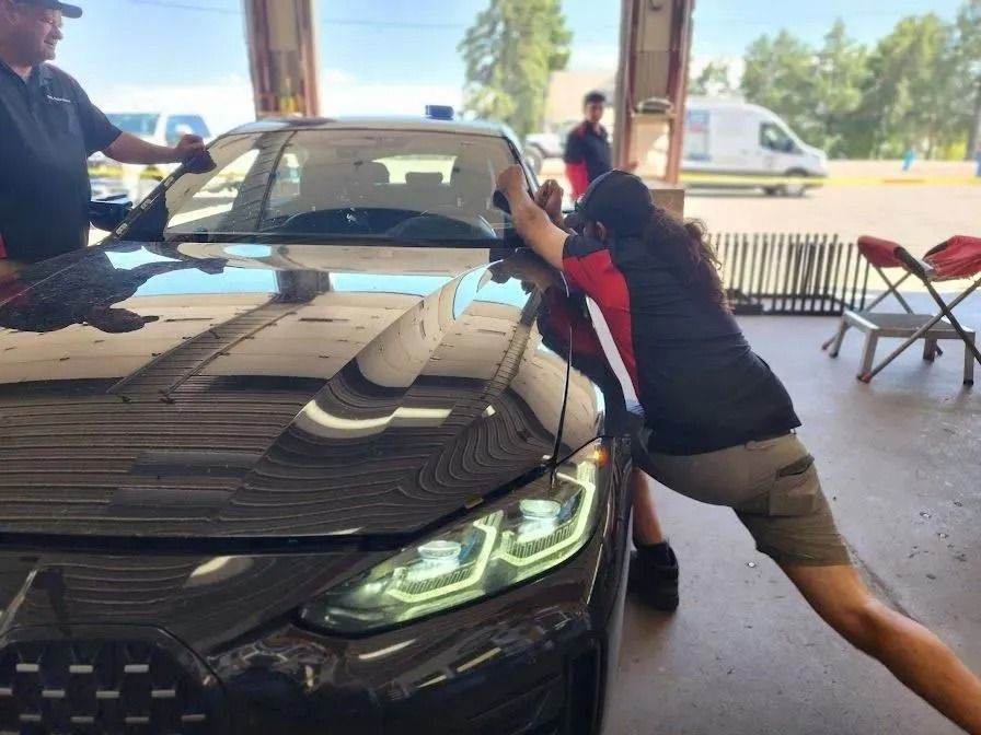 Two workers wearing uniforms apply a tinted film to the windshield of a black car parked inside an open-air garage.