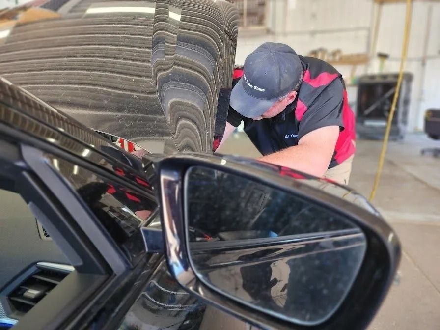 A mechanic in a red and black uniform leans over the open hood of a car inside a repair shop.