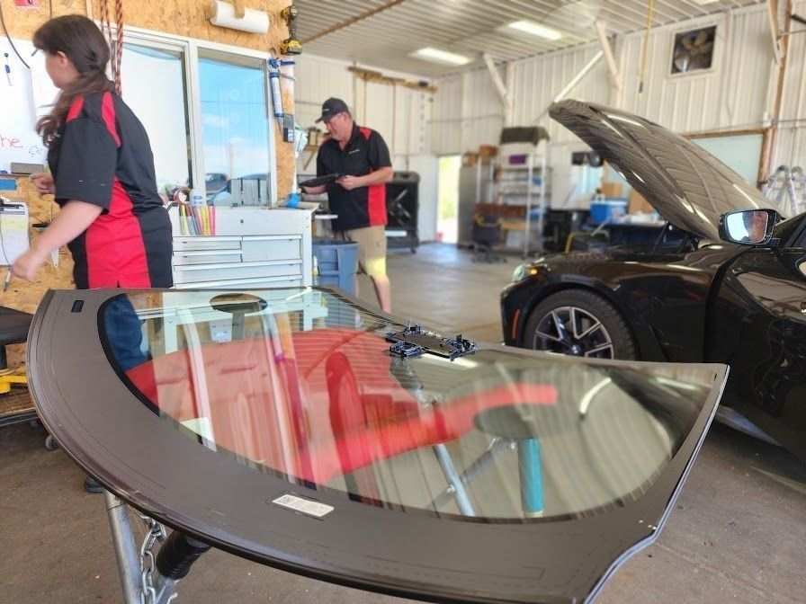 Two technicians in a garage prepare a new windshield, with a vehicle with its hood open in the background.