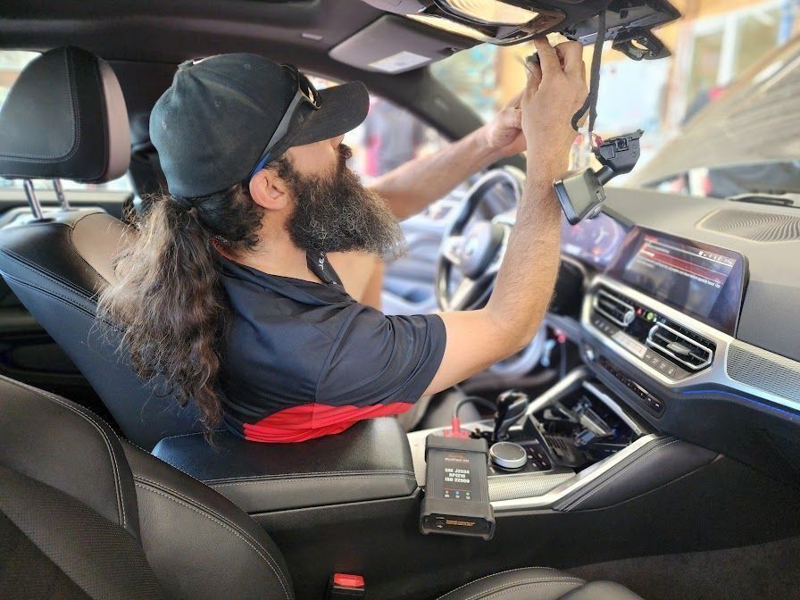 A person with a beard and ponytail sits in a car, installing a dashcam onto the rearview mirror.