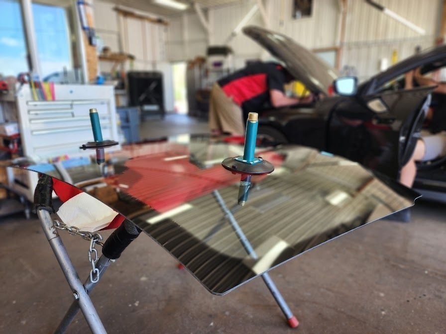 A car windshield with two suction cup handles sits on a stand in a garage, while a person works on a car in the background.