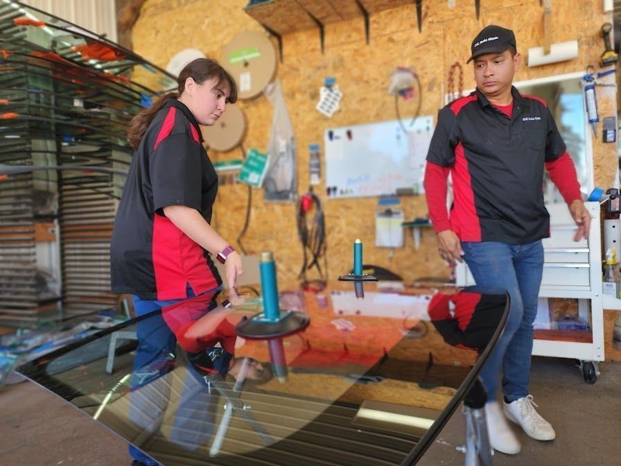 Two people in matching red and black uniforms work on a large piece of glass in a workshop.