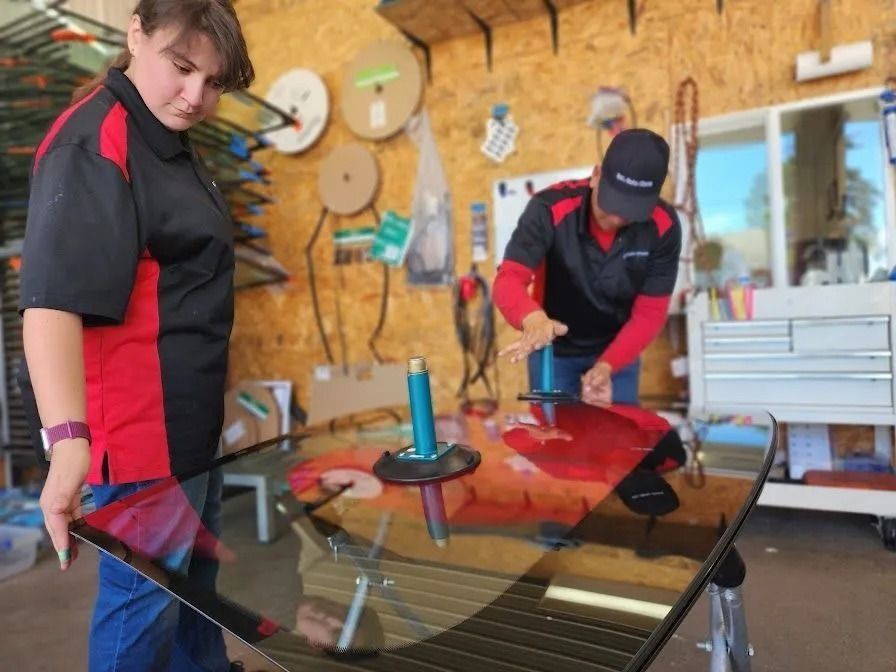 Two technicians in matching uniforms work together in a shop to position a large glass windshield using suction handles.