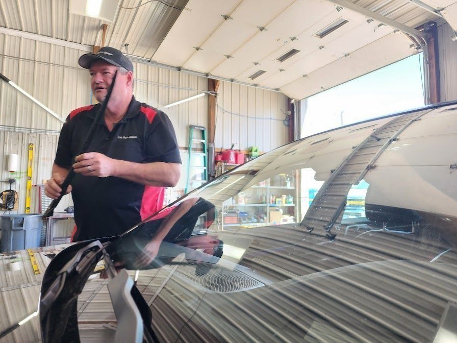 A person in a black and red polo shirt holds a windshield wiper blade while standing over a car in an auto repair shop.