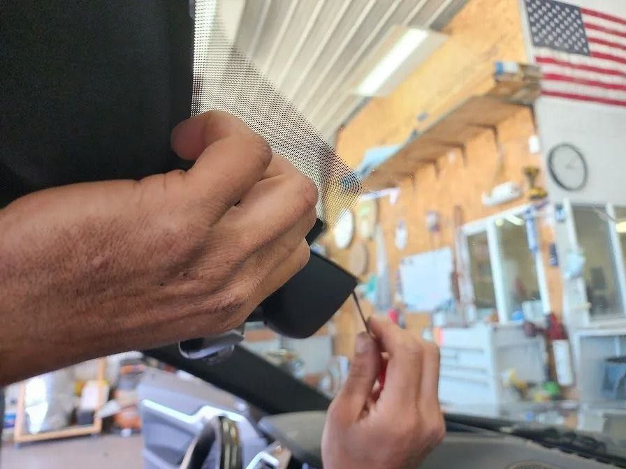 Hands working on the interior trim and rearview mirror area of a car windshield inside a garage.