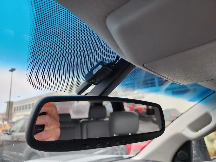 An interior view of a car windshield showing a rearview mirror, a black electronic device, and the black dot matrix.