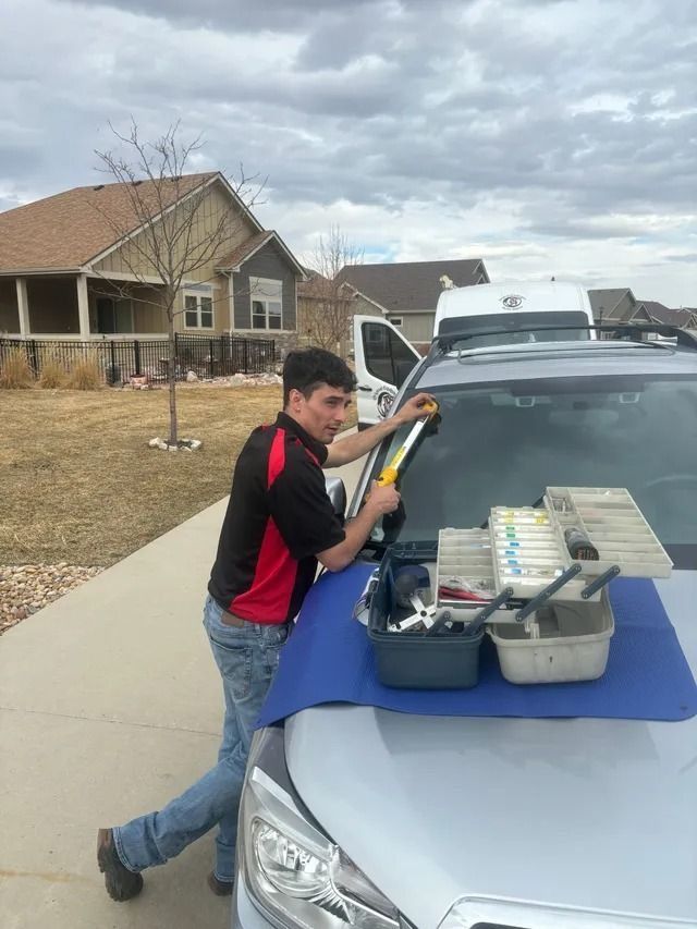 A technician in a red and black uniform repairs a car windshield, with a toolbox placed on the hood in a residential area.