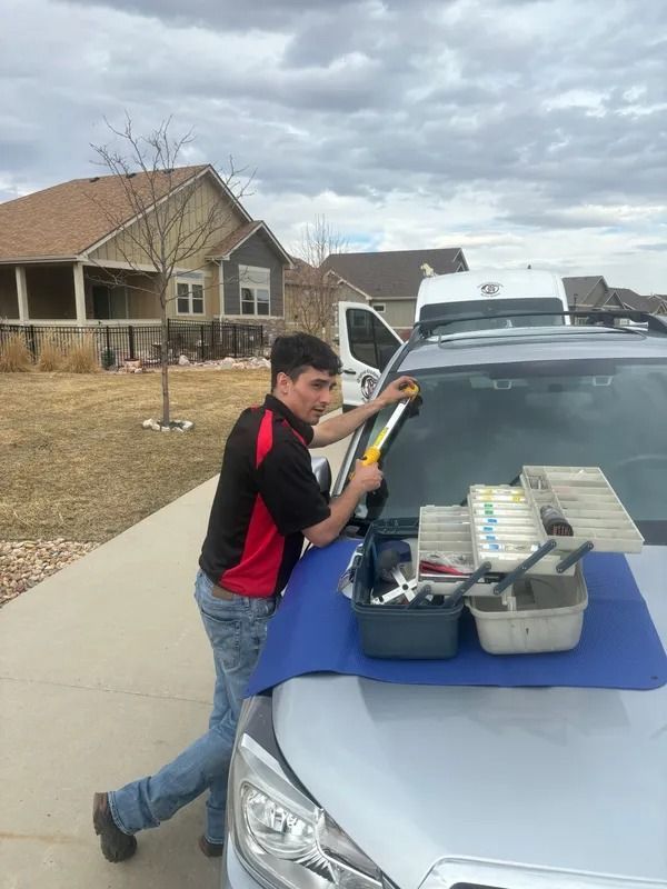Technician in a red and black shirt repairing a car windshield with tools in a residential driveway.