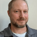 A man with a beard is wearing a gray shirt and smiling for the camera.