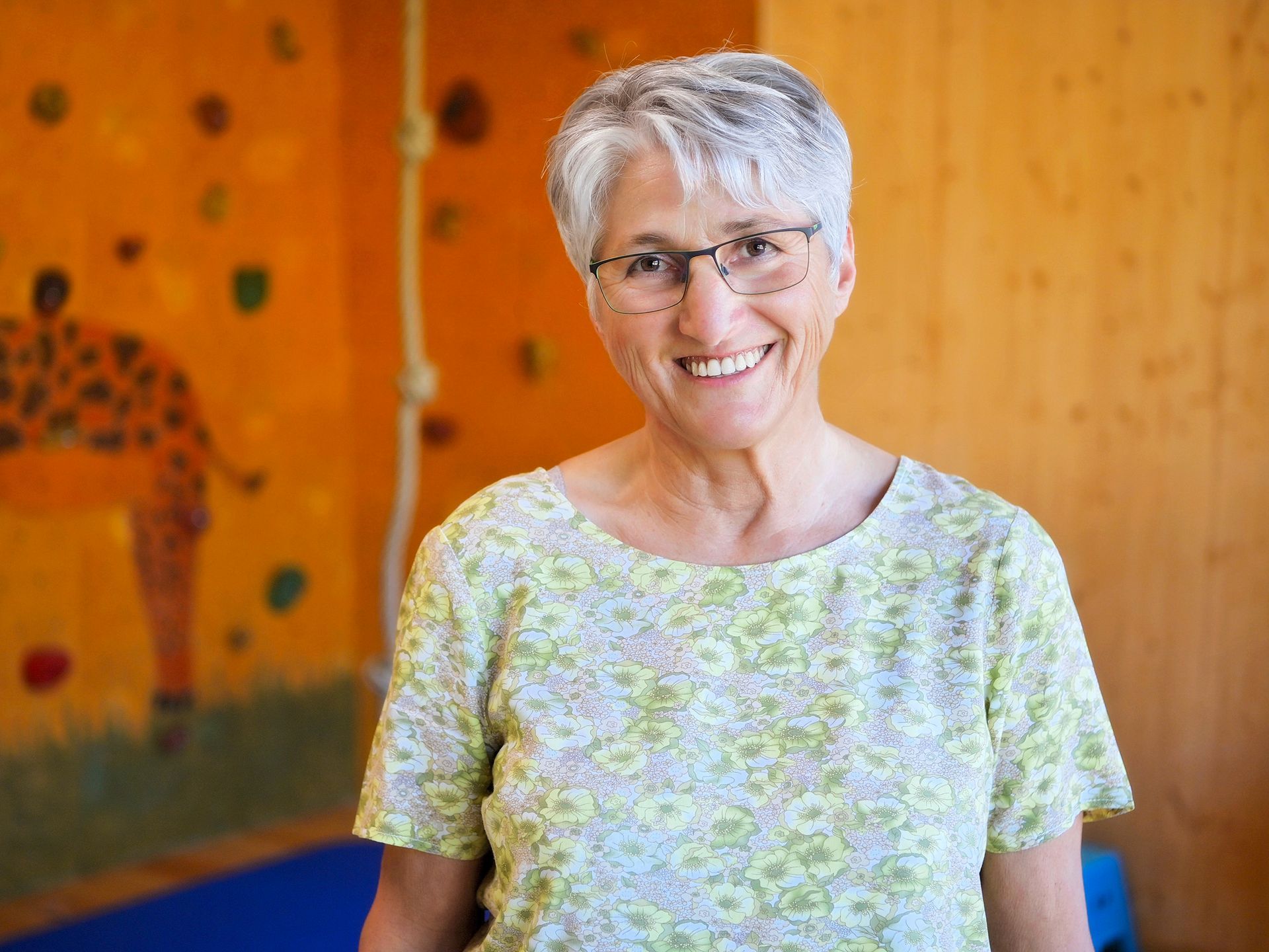 Eine Frau mit Brille lächelt vor einer Kletterwand.