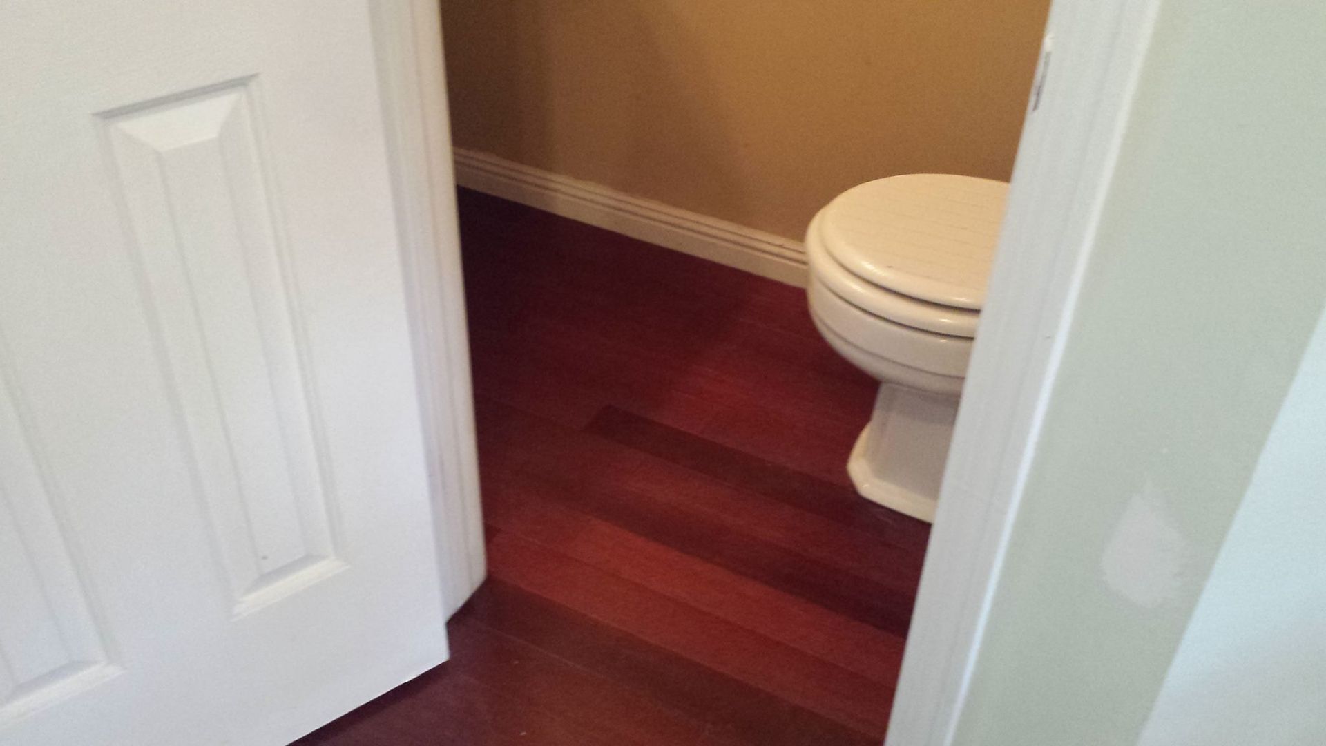 Partially open door revealing a small bathroom with a white toilet and red-toned wood floor.