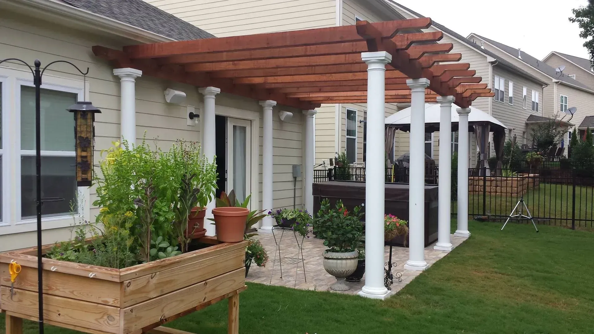 Pergola with white columns, attached to a house. Wooden beams, plants, and a lawn.
