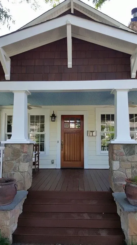 Front porch of a house with stone pillars, wooden door, steps, and windows. Brown and white exterior.