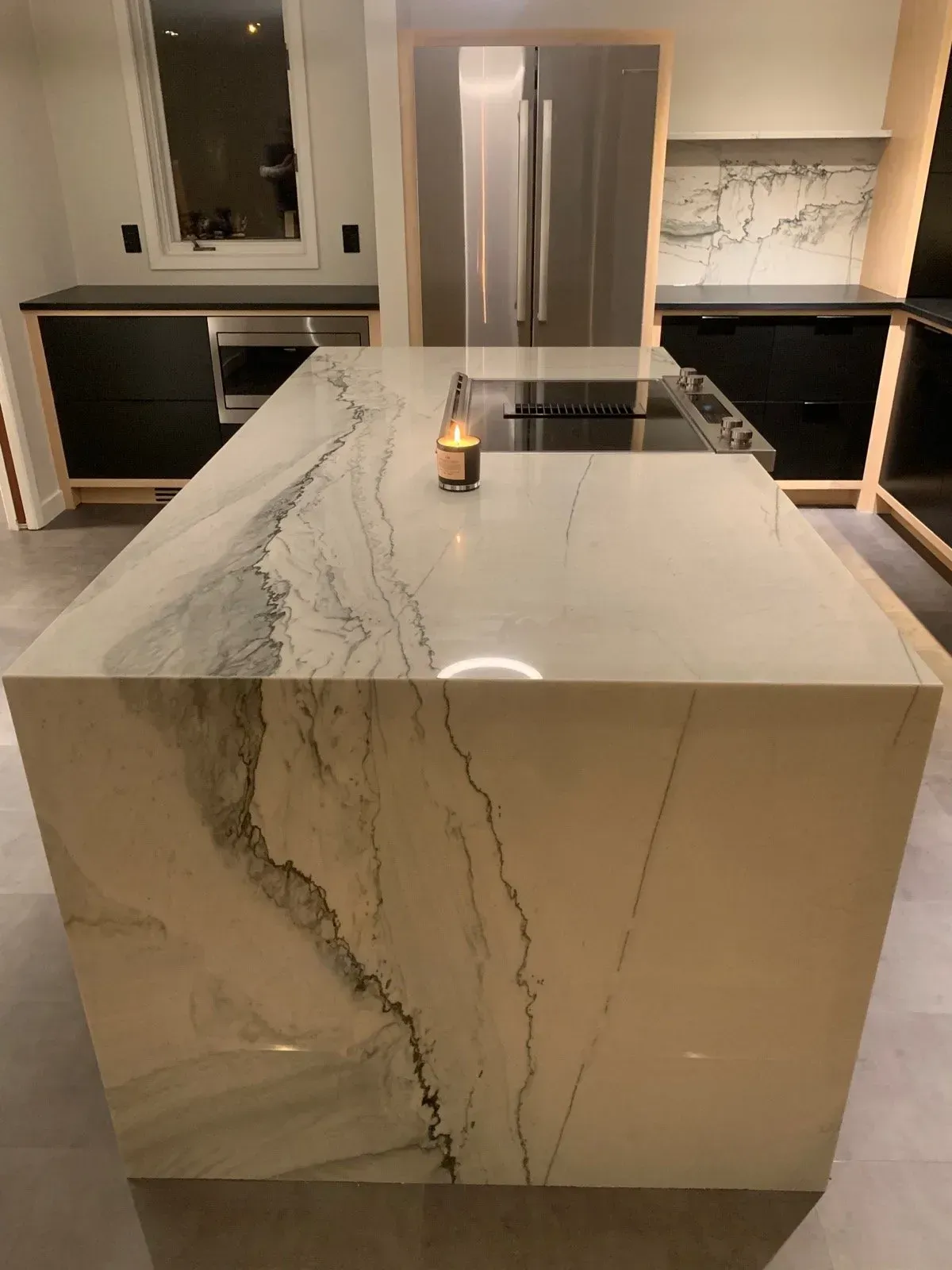 Modern kitchen with large marble island, stainless steel refrigerator, and dark cabinets.