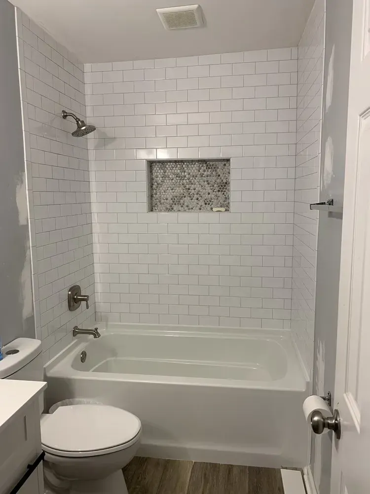 White-tiled bathroom with a bathtub, shower, and toilet. A niche with speckled tile is in the wall.