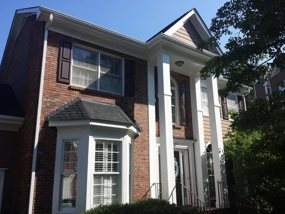 Two-story brick house with white pillars, brown shutters, and dark roof under a blue sky.