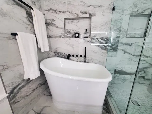 White freestanding bathtub in a marble-tiled bathroom with black fixtures and a glass shower door. White towels on a bar.
