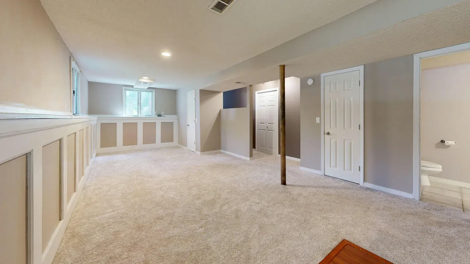 Empty basement with carpet, tan walls, wainscoting, a support beam, and a closed white door.