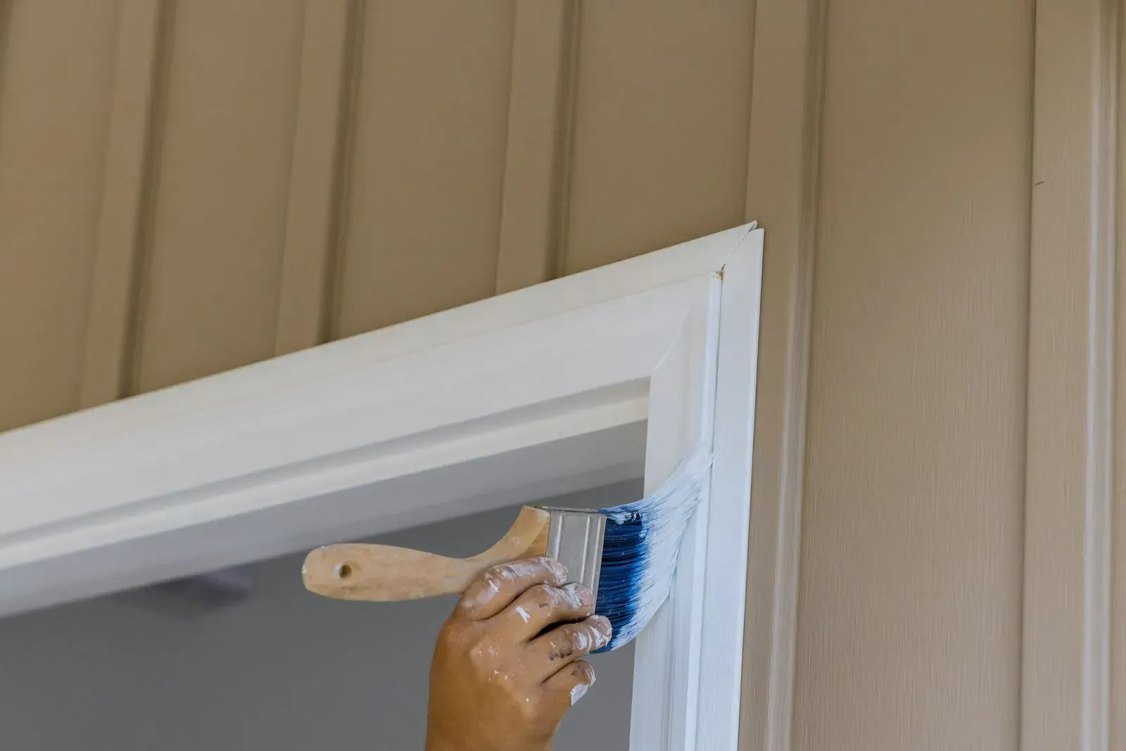 Hand painting white trim on a doorway with a brush; beige siding in background.