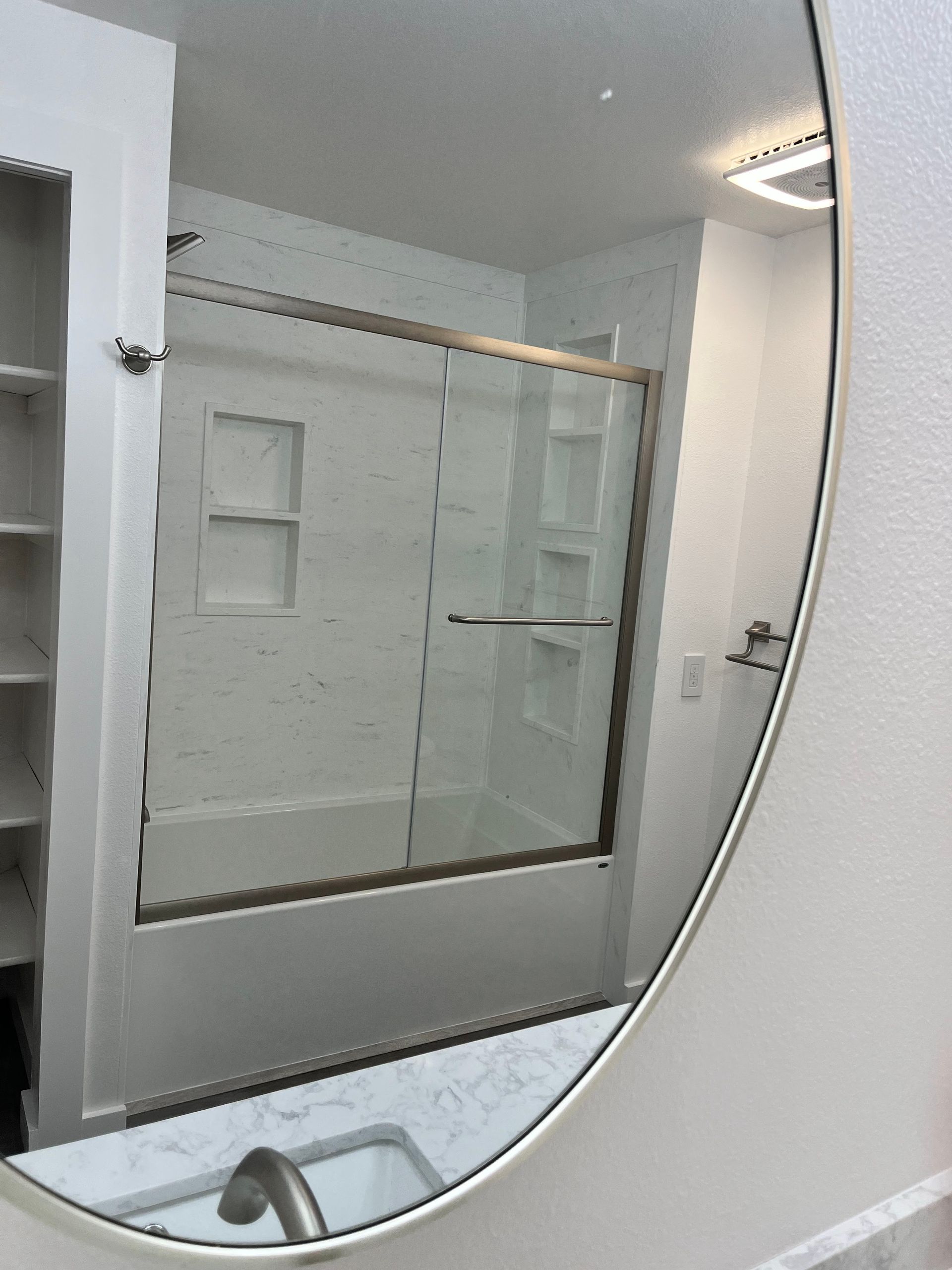 Oval mirror reflecting a white bathroom with a tub/shower, built-in shelves, and a marble countertop.