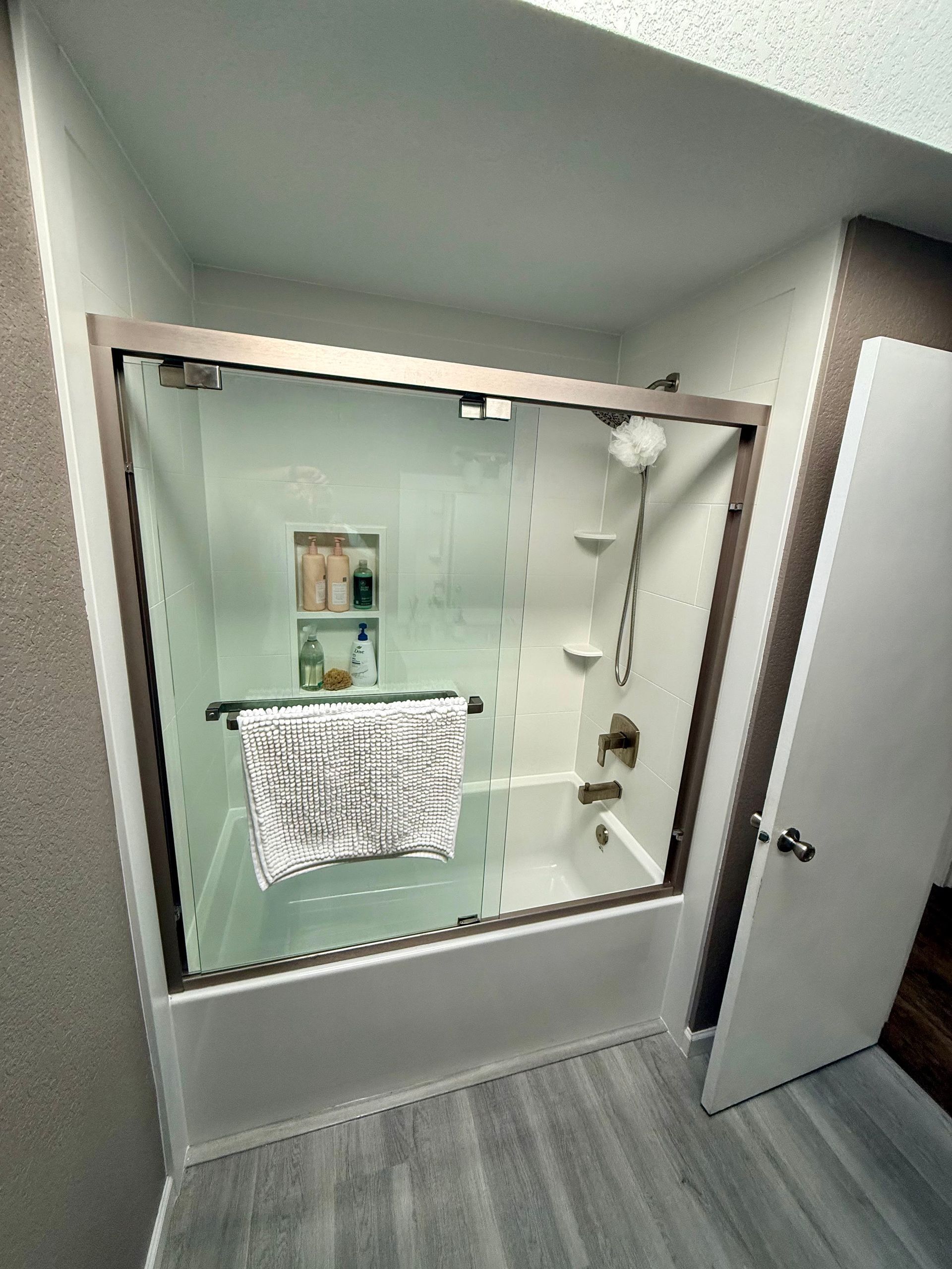 Bathroom with a glass shower door, white tub, and gray flooring. A towel hangs on a bar inside.