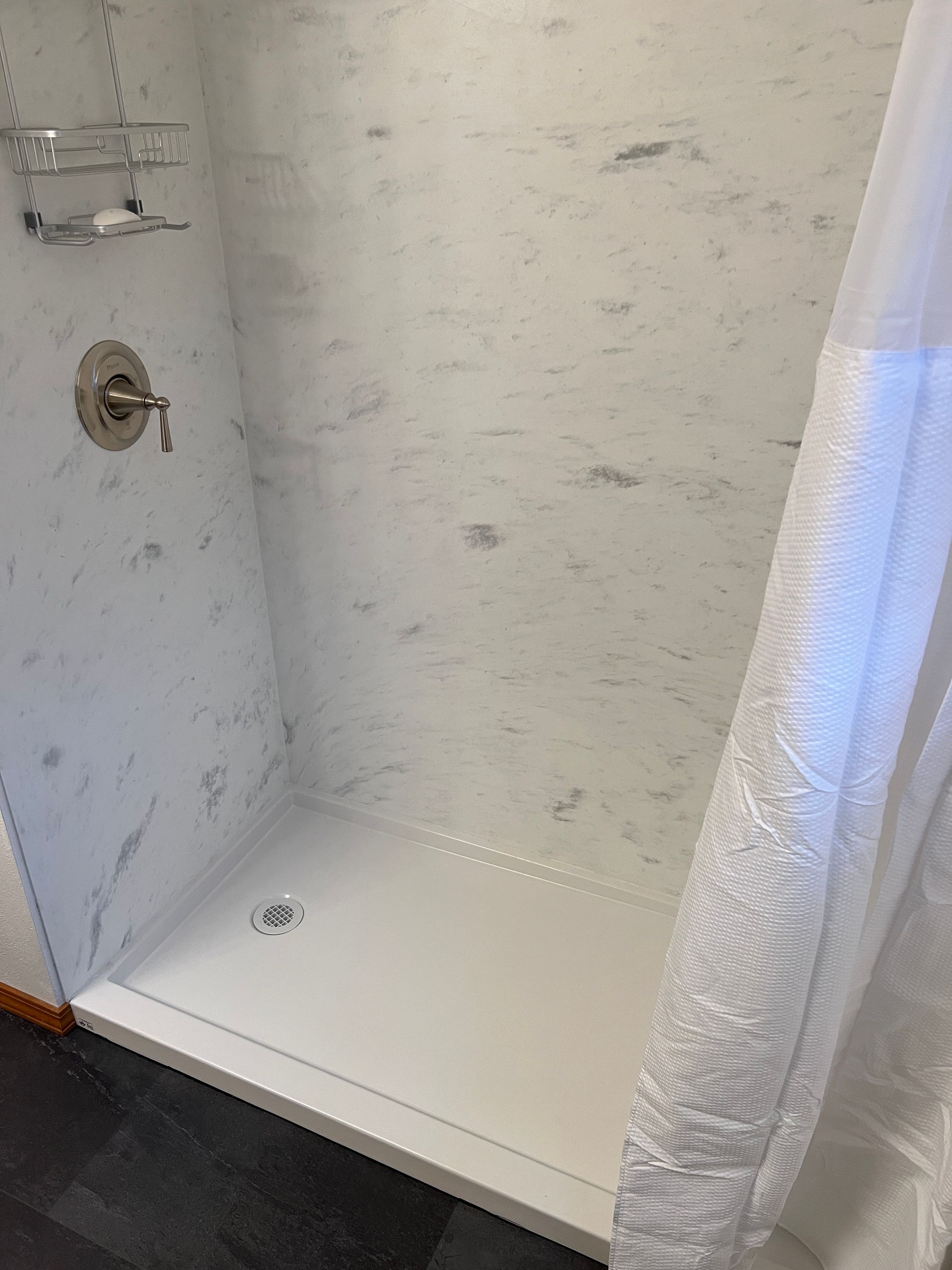 White shower with marble-like walls, a low threshold, and a silver showerhead; a white shower curtain hangs on the right.