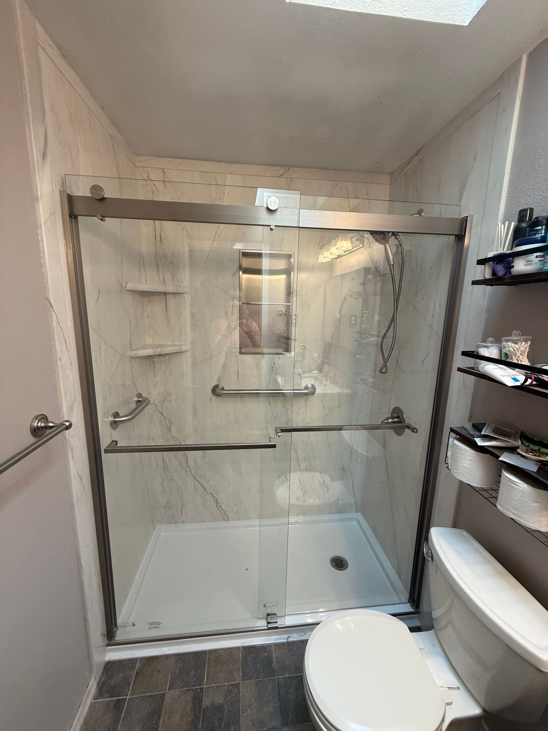 Accessible shower with grab bars and a sliding glass door, next to a toilet and shelves.