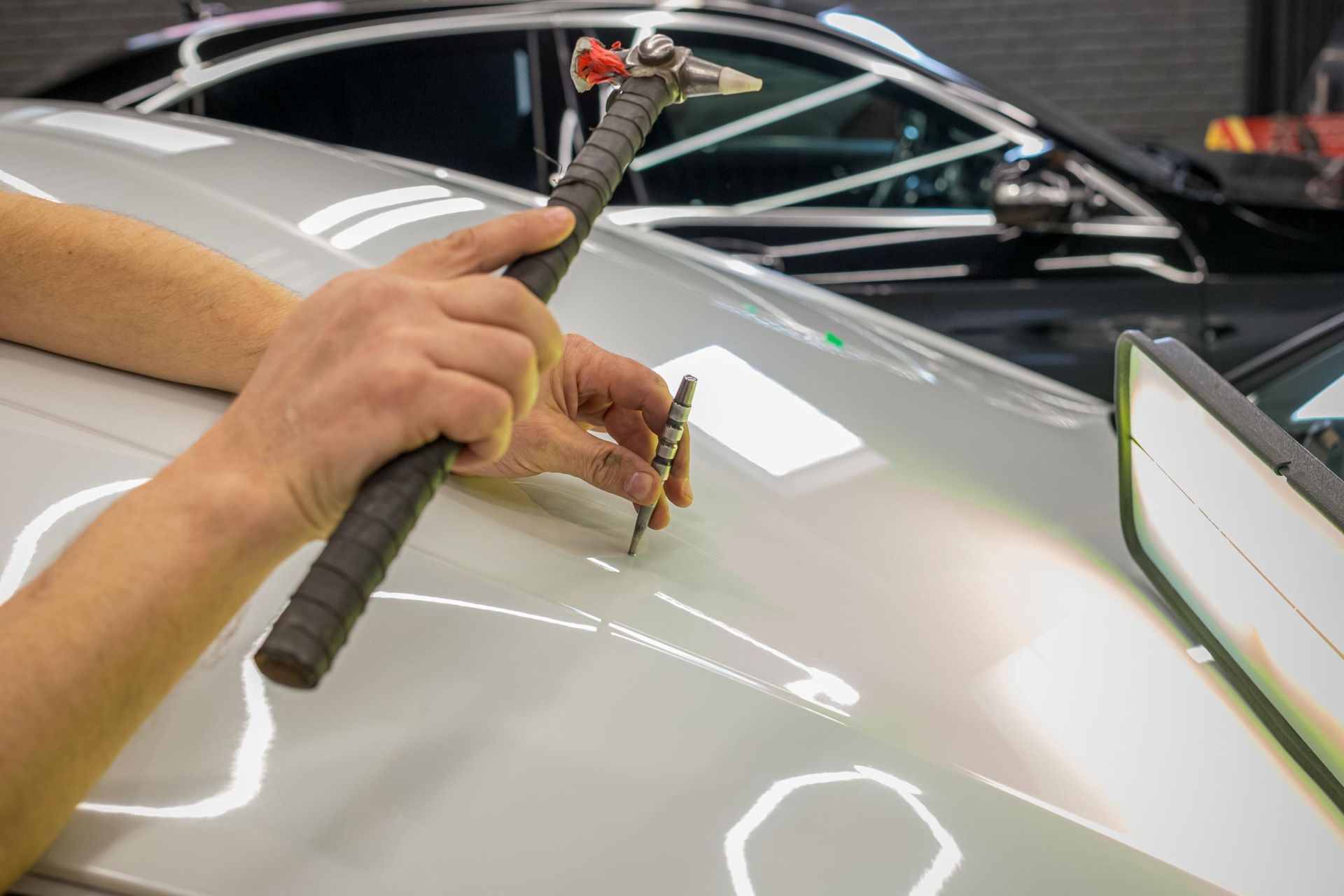 Hand repairing a car’s white hood with a tool in an auto shop