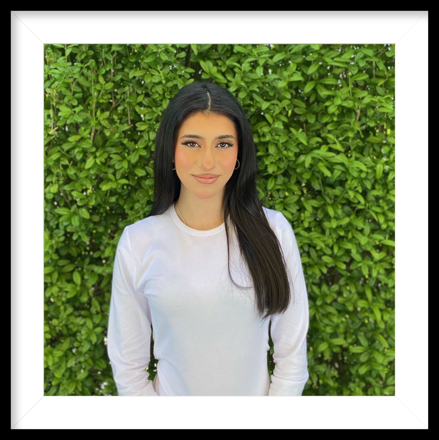 Eine Frau in einem weißen Hemd steht vor einer grünen Hecke.