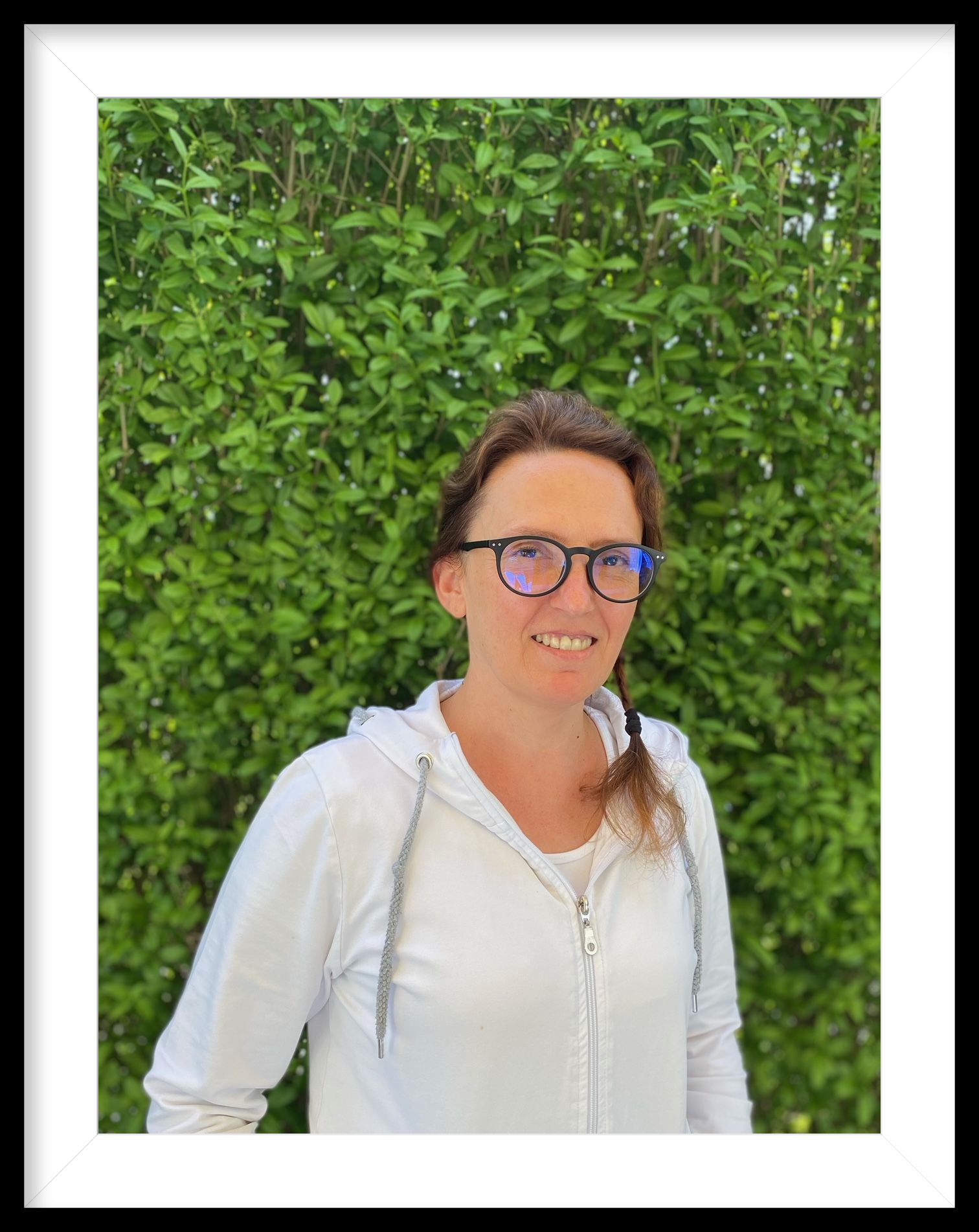 Eine Frau mit Brille und weißem Kapuzenpullover steht vor einer grünen Hecke.