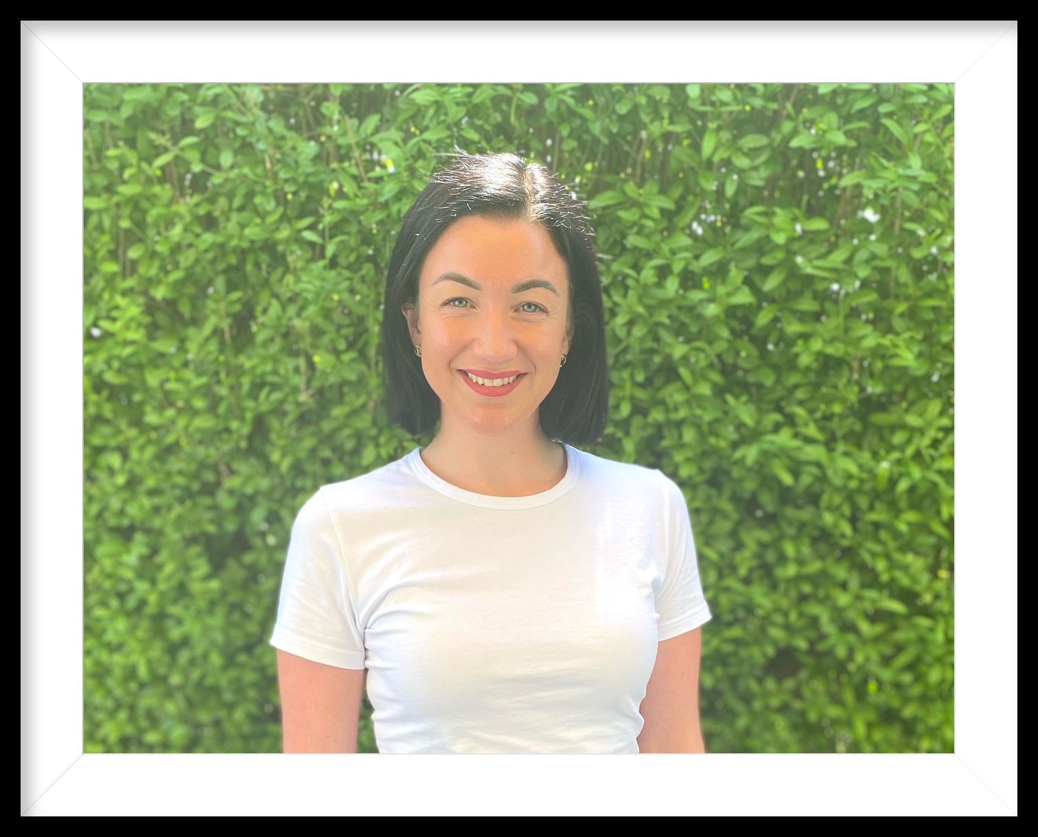 Eine Frau in einem weißen Hemd lächelt vor einer grünen Hecke.