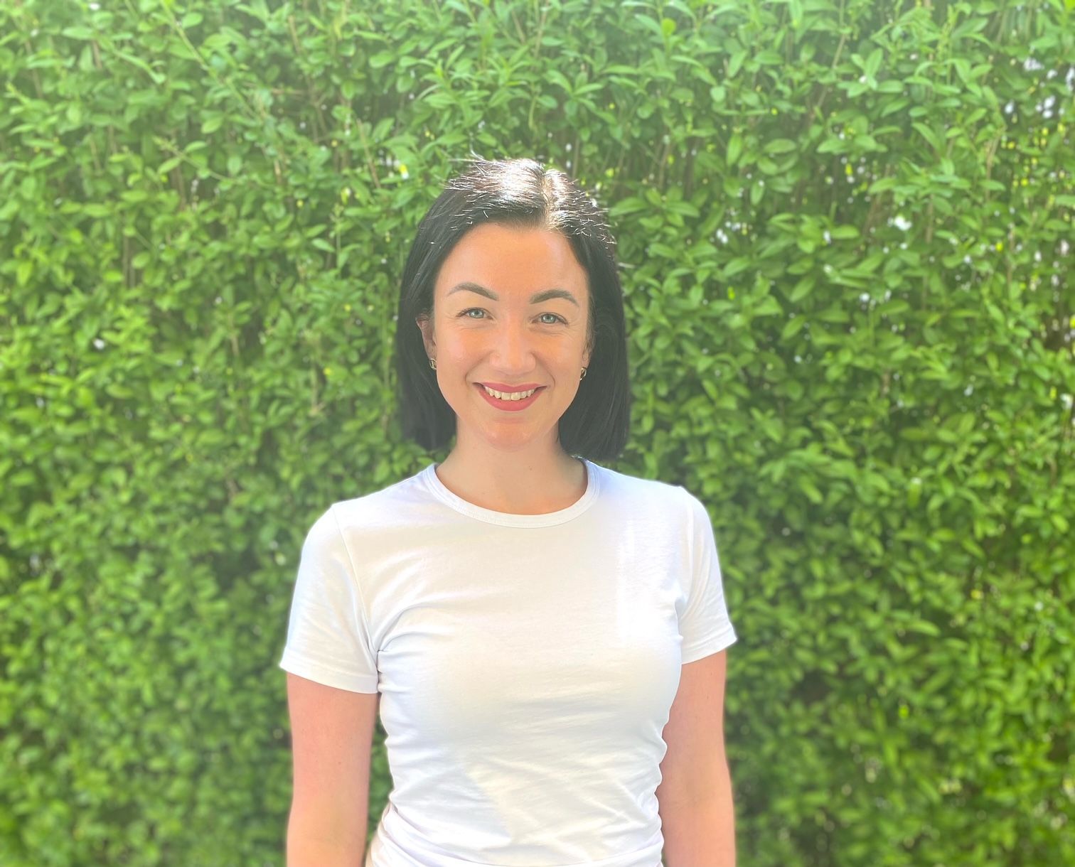 Eine Frau in einem weißen T-Shirt lächelt vor einer grünen Hecke.