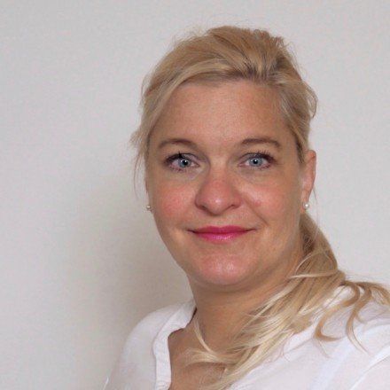 Eine Frau mit blonden Haaren und blauen Augen lächelt in die Kamera.