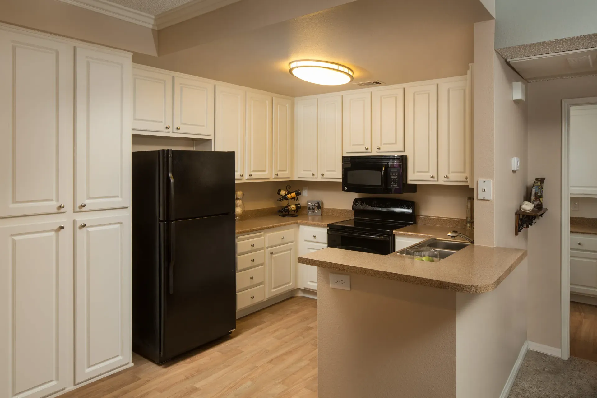 Open apartment kitchen with white cabinets, beige countertops, and black appliances.
