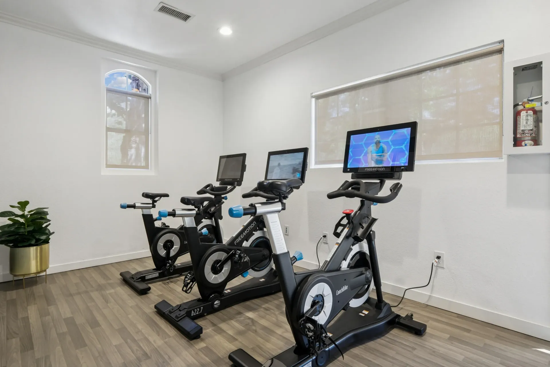 Light-filled community gym with three stationary bikes and wall-mounted screens.