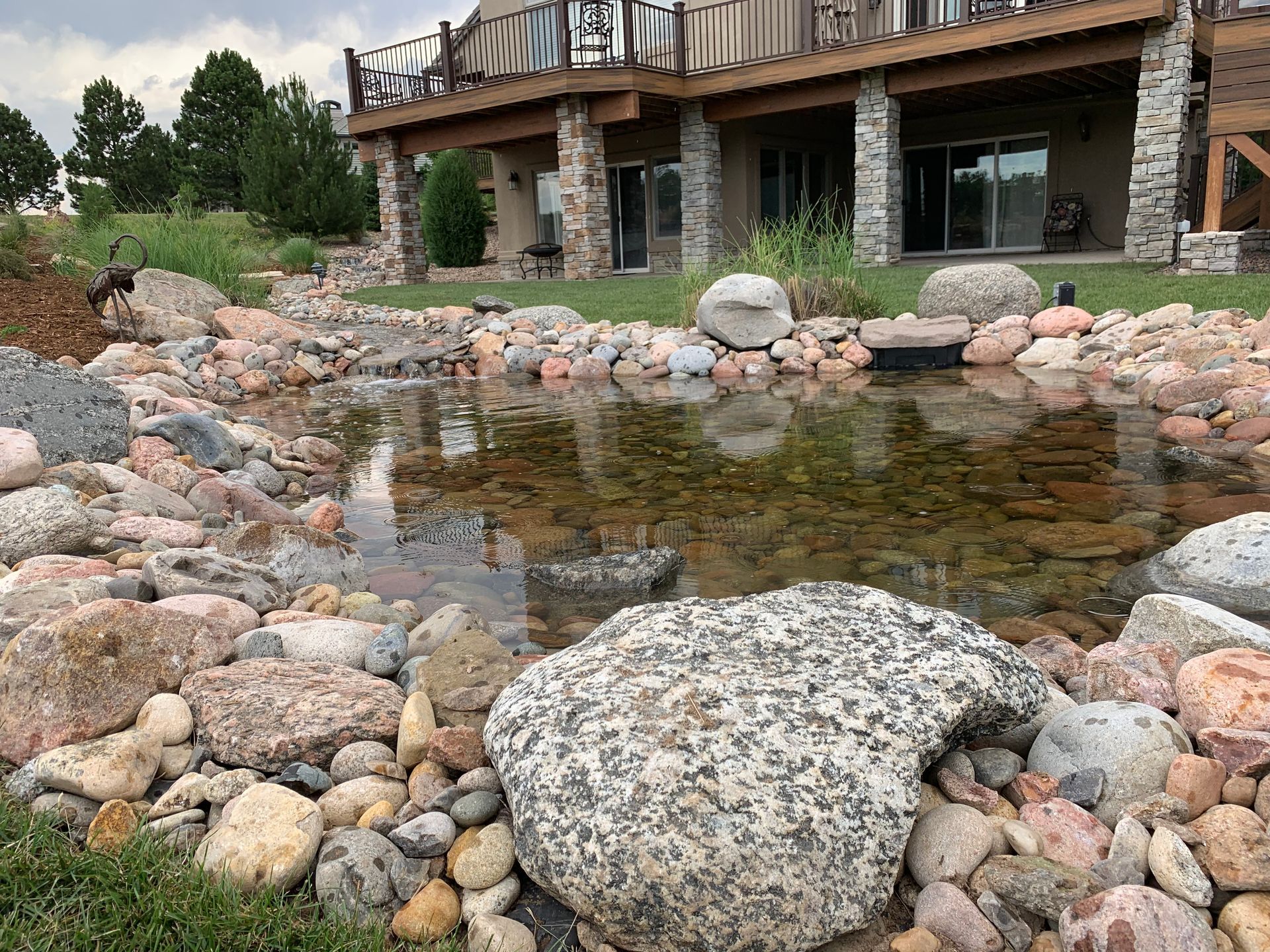 Fountain with tiered basins, water spraying upwards. Green trees, wooden fence and building in the background.