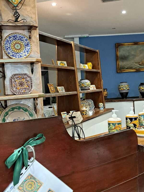 Interno di un negozio con scaffali che espongono piatti e vasi in ceramica. Panca in legno in primo piano. Parete blu.