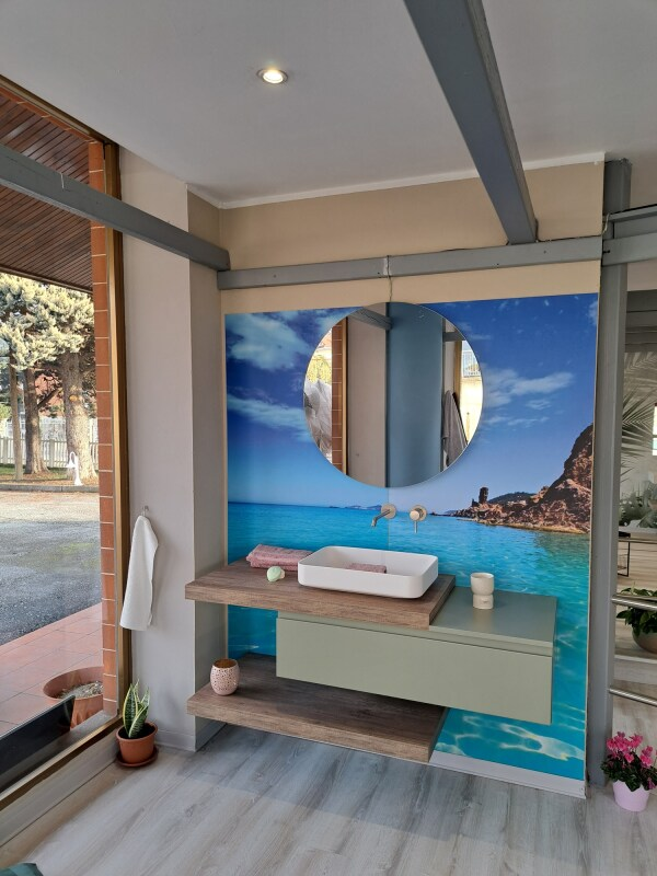 Bagno con lavandino, mobiletto e specchio rotondo su sfondo murale raffigurante il mare; legno chiaro e colori blu-verde.