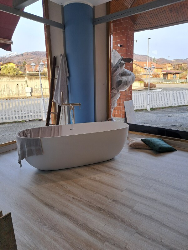 Vasca da bagno ovale bianca in una stanza luminosa con pavimento effetto legno chiaro. Dietro si trovano una colonna blu e una finestra.