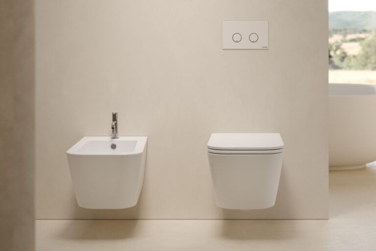Bidet e WC bianchi sospesi, uno accanto all'altro. Comando di scarico sopra il WC. Bagno dai toni neutri.