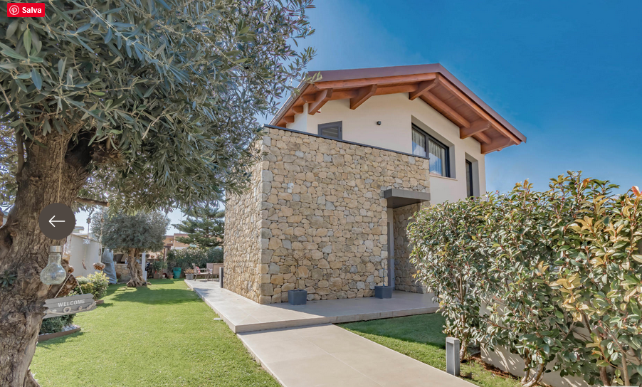 Casa con muro in pietra, tetto in legno, immersa nel verde. Giornata di sole.