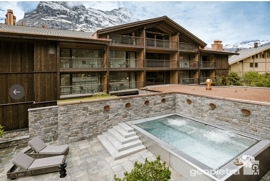 Esterno dell'hotel con piscina, montagne sullo sfondo, facciata in legno, pietra grigia, due sedie a sdraio.