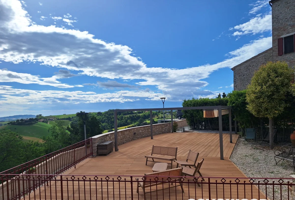 Terrazza in legno arredata con vista sulla valle sotto un cielo azzurro e nuvoloso.