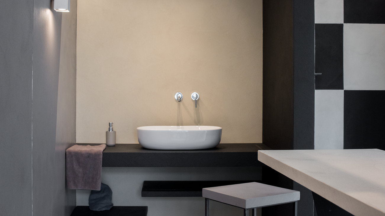 Bagno con lavandino ovale bianco su piano di lavoro nero, parete beige, piastrelle a scacchi bianche e nere.