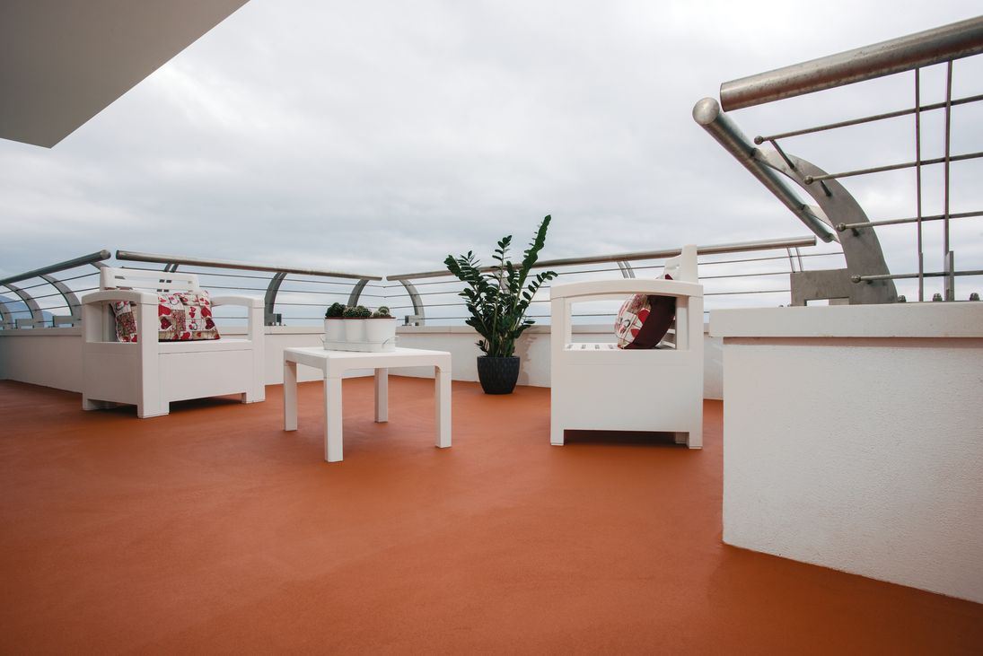 Patio sul tetto con mobili bianchi, piante e ringhiera in vetro contro un cielo nuvoloso.