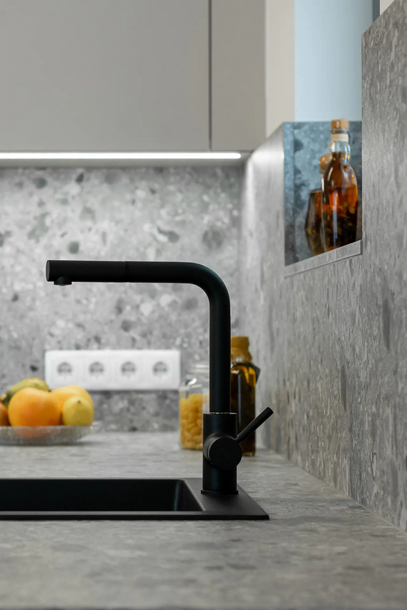 Rubinetto nero della cucina sopra un lavandino scuro, contro un piano di lavoro e un paraschizzi grigi maculati.